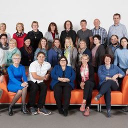 Gruppenfoto eines Teams von Fachleuten in Geschäftskleidung, die auf orangefarbenen Sofas vor einem weißen Hintergrund stehen und sitzen.
