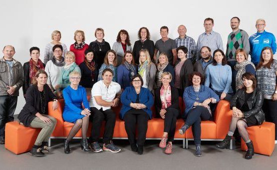 Gruppenfoto eines Teams von Fachleuten in Geschäftskleidung, die auf orangefarbenen Sofas vor einem weißen Hintergrund stehen und sitzen.