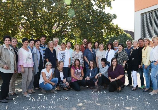 Eine Gruppe von Erwachsenen posiert für ein Foto vor einem Gebäude mit Bäumen, einige lächeln, andere schauen in die Kamera, mathematische Gleichungen sind auf dem Boden gezeichnet.
