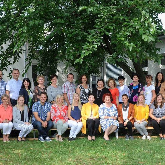 Eine Gruppe von Menschen posiert für ein Foto in einem Garten mit einem Baum, stehend und sitzend auf einer Bank. Hinter ihnen steht ein weißes Gebäude mit Glasfenstern.