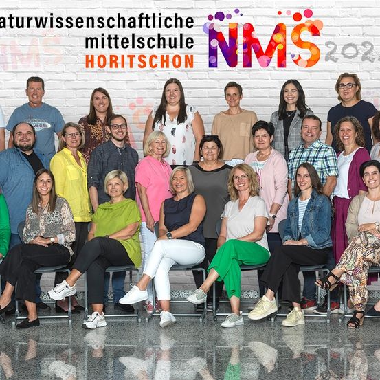 Eine Gruppe von Menschen sitzt vor einer Ziegelwand mit dem Text 'Naturwissenschaftliche Mittelschule Horitschon NMS 2021' über ihnen. Sie tragen alle Freizeitkleidung.