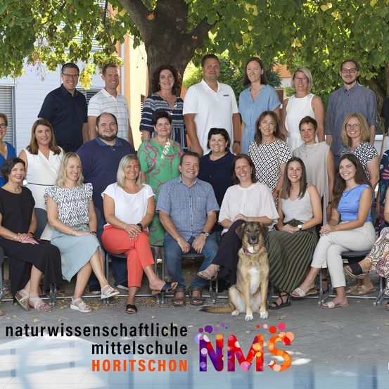 Ein Gruppenfoto von Menschen, einschließlich Erwachsener und eines Hundes, die unter einem Baum sitzen. Sie sind wahrscheinlich Mitarbeiter oder Fakultätsmitglieder einer Schule. Der Name der Schule ist Horitschon.
