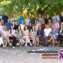Eine Gruppe Erwachsener posiert für ein Foto unter einem Baum an einer Schule. Sie alle lächeln und tragen Freizeitkleidung. Das Bild hat einen Wasserzeichen mit dem Text 'naturwissenschaftliche mittelschule HORITSCHON'.