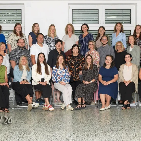 Eine Gruppe von Menschen, sowohl Männer als auch Frauen, sitzen und stehen in einem Raum, wahrscheinlich für ein Gruppenfoto. Sie sind in verschiedenen Outfits gekleidet.