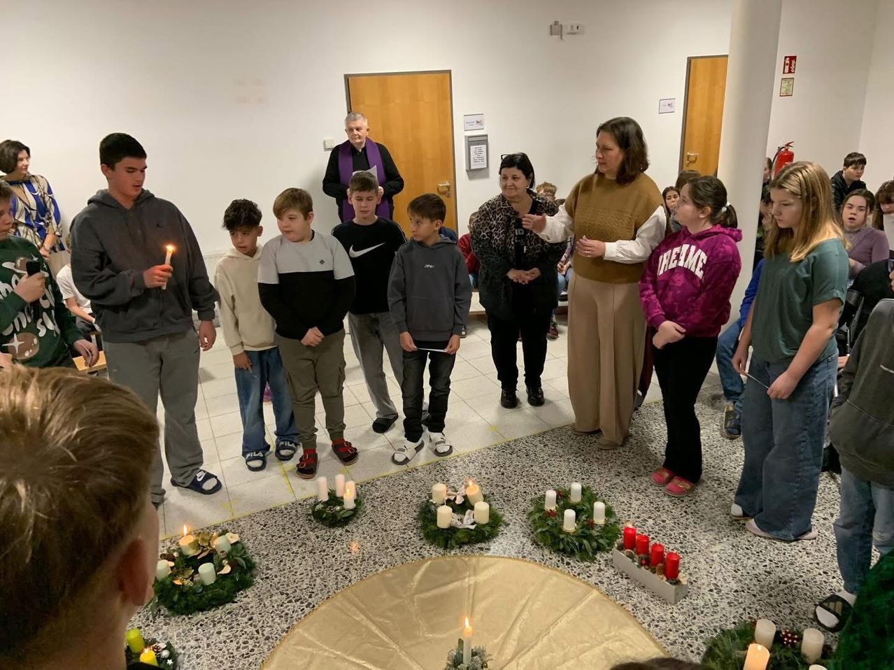 Eine Gruppe von Kindern und Erwachsenen steht um eine Kerzengirlande herum. Ein Erwachsener hält eine brennende Kerze, während andere in der Nähe stehen. Der Raum hat weiße Wände und Holztüren.