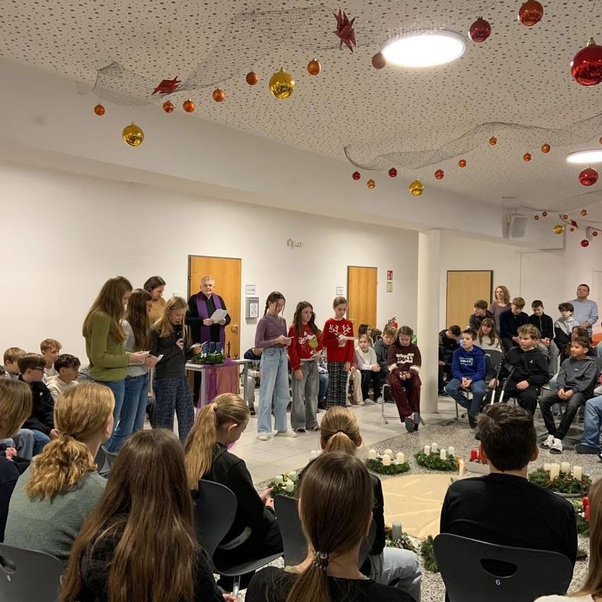 Eine Gruppe von Kindern und Erwachsenen versammelt sich in einem Raum für eine Veranstaltung. Sie sitzen auf Stühlen und stehen um einen Kreis von Kerzen herum. Einige halten Papiere. Der Raum ist mit Weihnachtskugeln und Lichtern geschmückt.