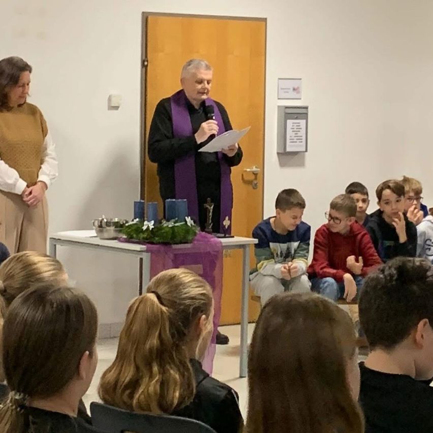 Ein Priester in einer violetten Stola spricht in ein Mikrofon an einem Tisch mit Kerze und Kreuz in einem Raum mit Kindern und einer Frau.