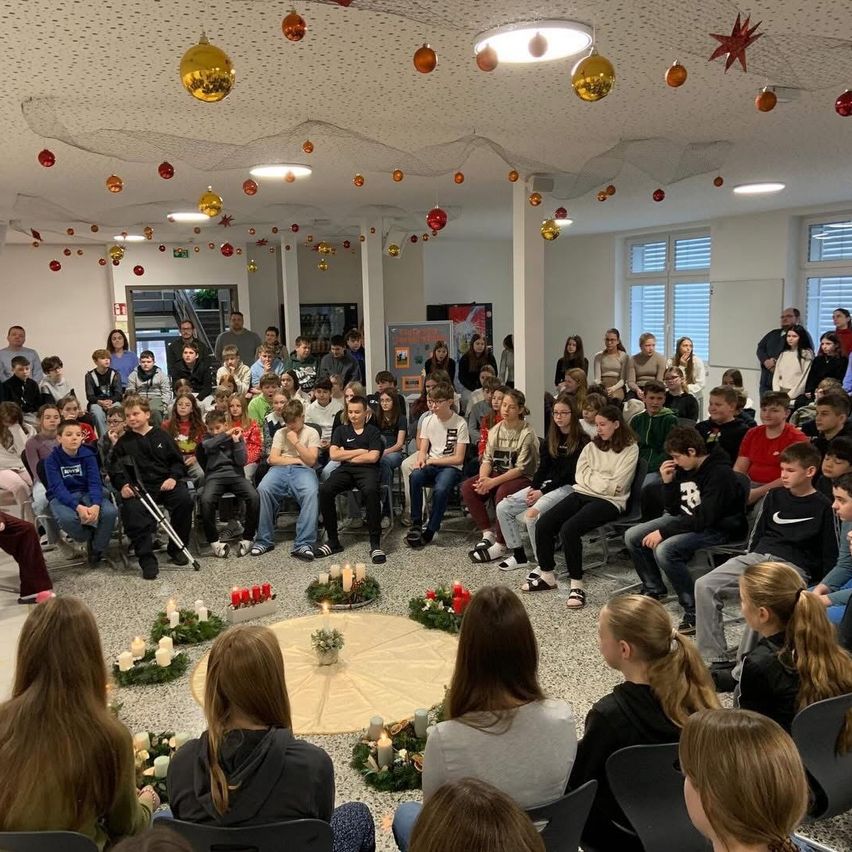 Eine Gruppe von Menschen in einem Raum mit brennenden Kerzen und Weihnachtsdekoration an der Decke. Einige Leute sitzen, andere stehen.