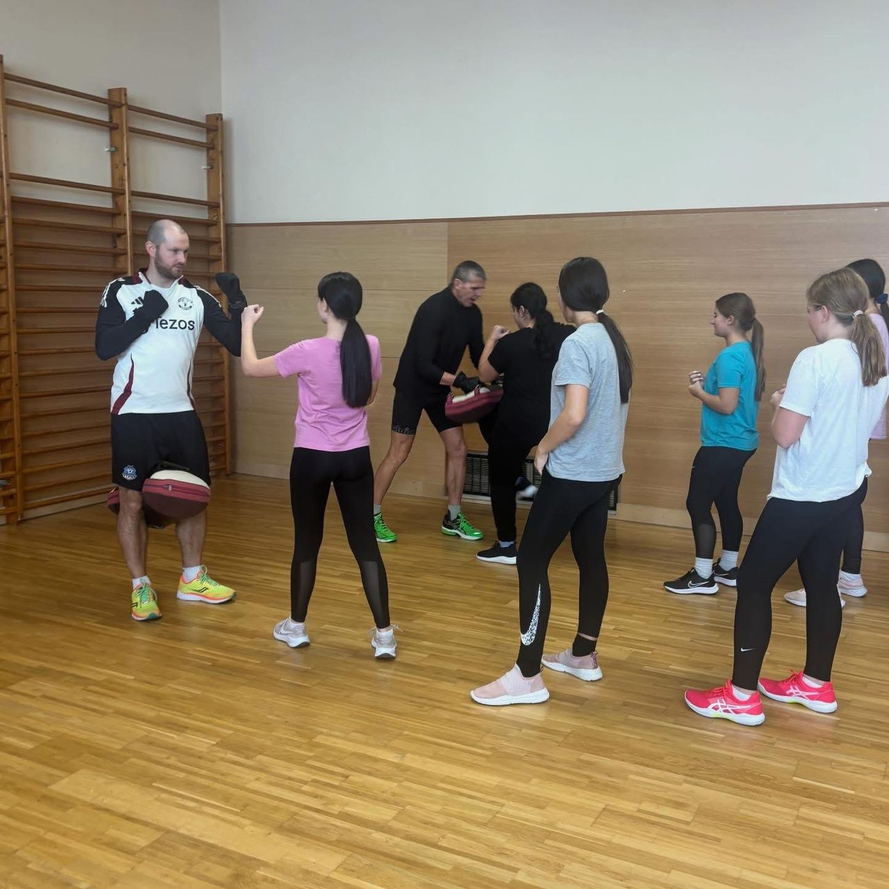 Eine Gruppe von Menschen trainiert Boxen in einem Fitnessstudio, wobei ein Mann einen Ball hält und ein Manchester United-Trikot trägt.