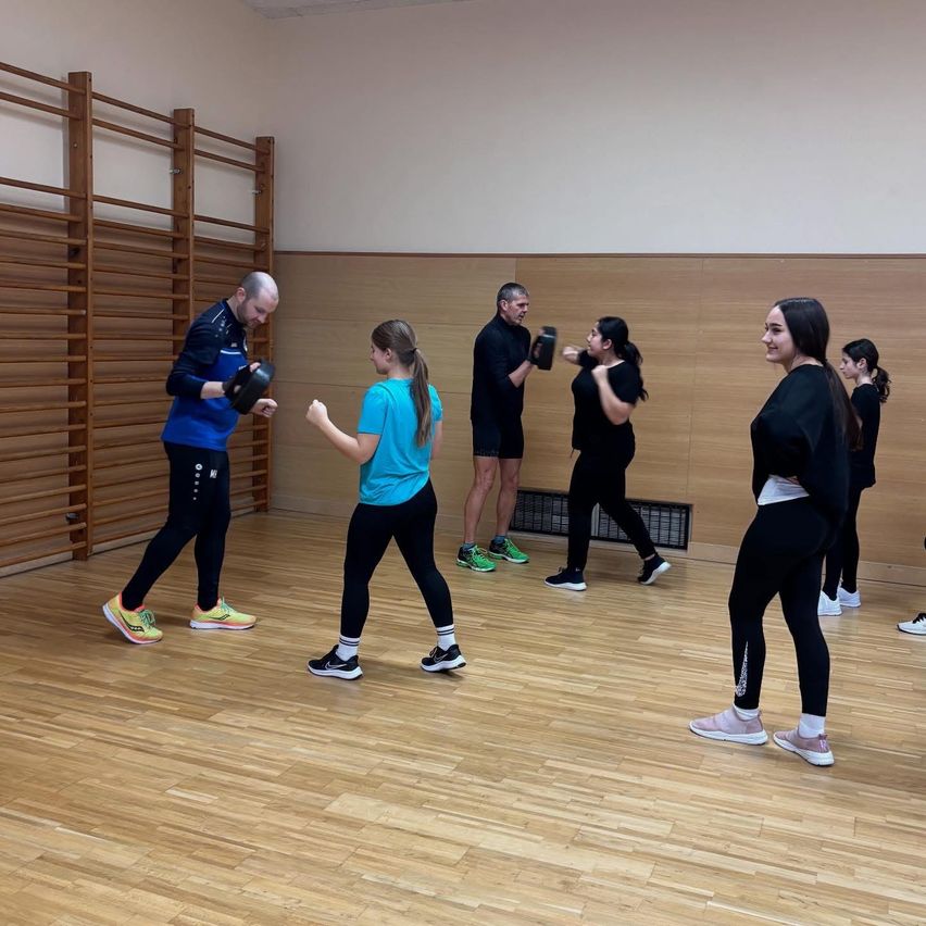 Eine Gruppe von Menschen befindet sich in einem Fitnessstudio. Einige benutzen Boxhandschuhe, während andere das nicht tun. Sie tragen Sportbekleidung und Turnschuhe. Hinter ihnen steht eine Holzwand mit Ausrüstung.