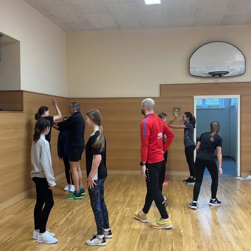 Eine Gruppe von Menschen versammelt sich in einem Raum, möglicherweise für einen Fitnesskurs. Sie tragen Sportbekleidung. Ein Mann in einem roten Langarmshirt leitet den Kurs.