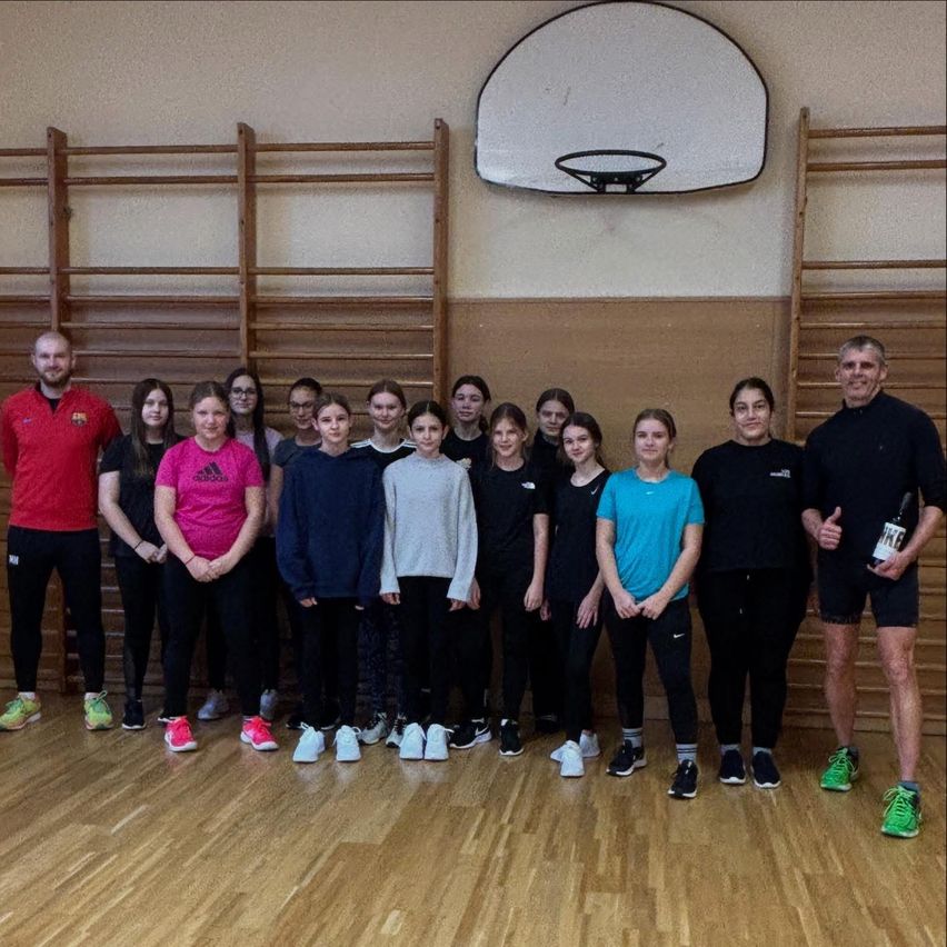 Eine Gruppe von Menschen, darunter zwei Männer, posiert für ein Foto in einer Turnhalle. Sie tragen Turnschuhe und einige tragen Brillen. Der Mann auf der rechten Seite hält eine Flasche. Hinter ihnen steht ein Basketballkorb.
