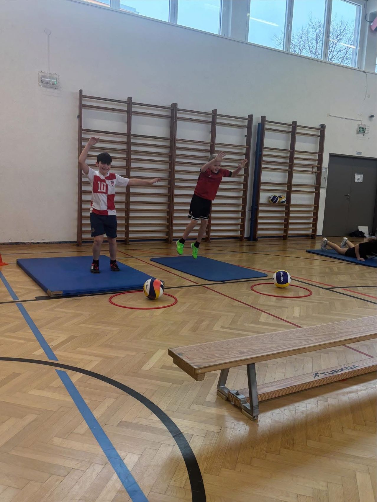 Zwei Jungen spielen ein Spiel in einer Turnhalle mit Bällen. Ein Junge in Rot springt, während der andere in Weiß zum Kicken bereit ist.