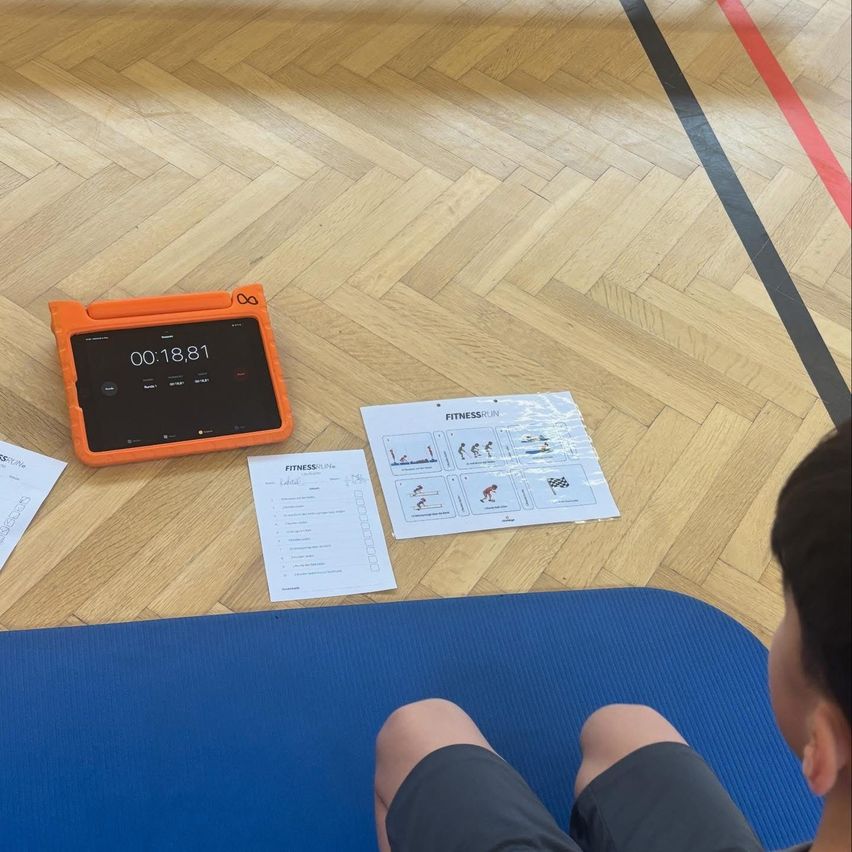 Ein Kind sitzt auf einer blauen Matte auf einem Holzboden und schaut auf ein Tablet mit der Zeit 00:18:81. In der Nähe liegen zwei bedruckte Papiere mit Übungen.