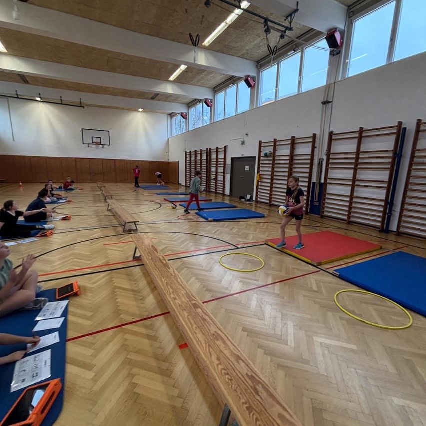 In einer Turnhalle sind mehrere Kinder bei verschiedenen Aktivitäten. Einige spielen mit Bällen und Hula-Hoops, während andere auf Matten sitzen und Übungen machen. Die Halle ist gut beleuchtet mit Holzböden und großen Fenstern.