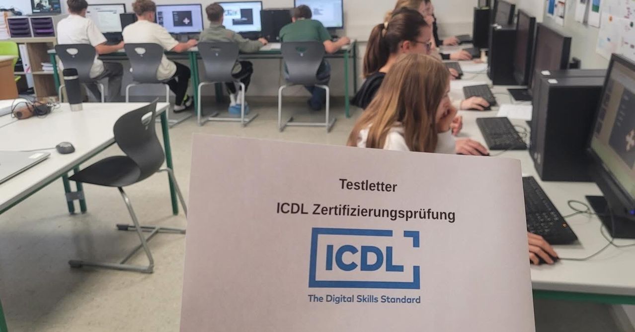 Ein Klassenzimmer mit Schülern, die an Computern arbeiten. Ein Testbrief für die ICDL-Zertifizierungsprüfung wird vor der Kamera gehalten.