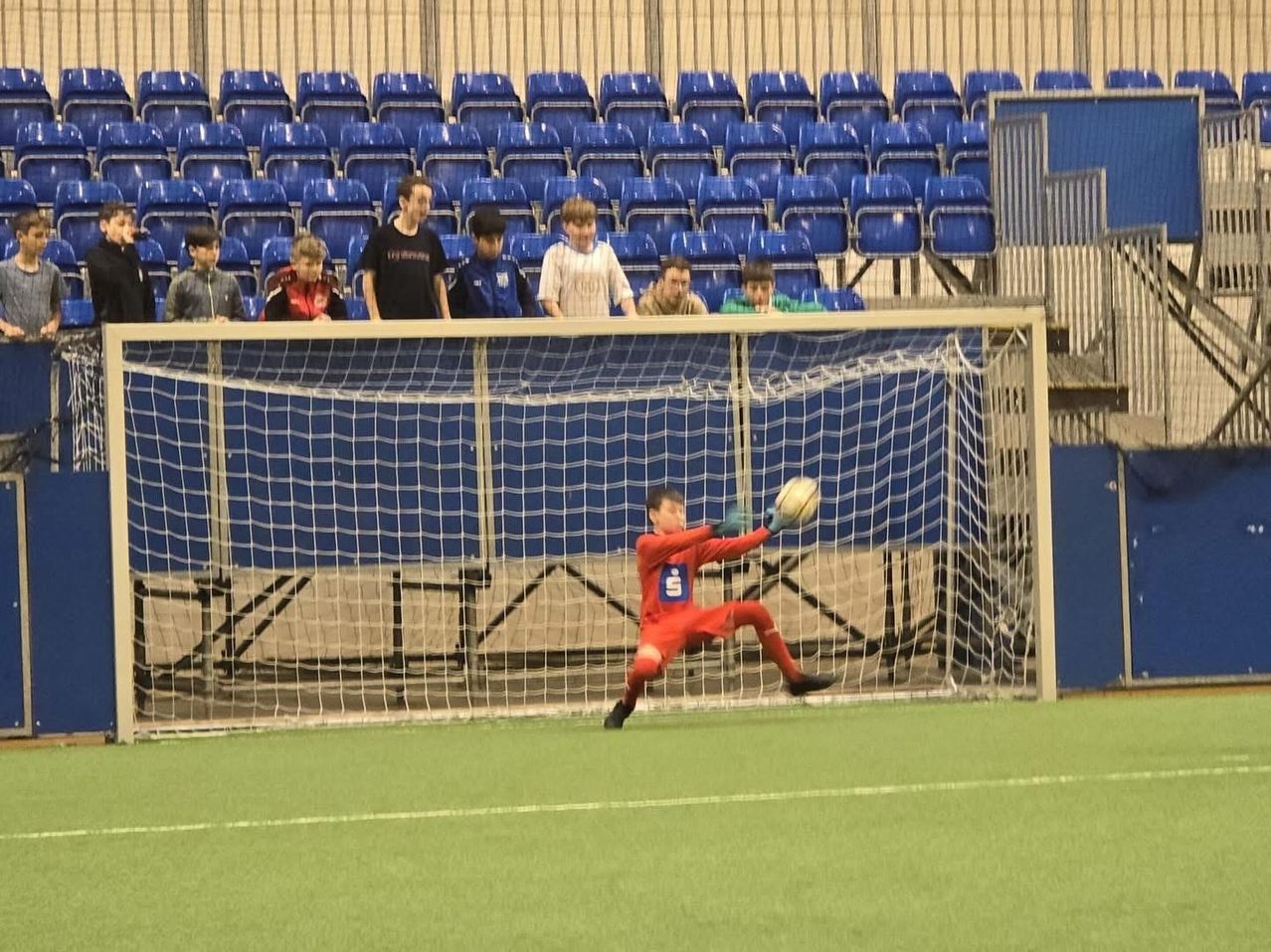 Ein Fußballtorwart taucht, um den Ball zu fangen, während mehrere Zuschauer von den Tribünen zusehen.