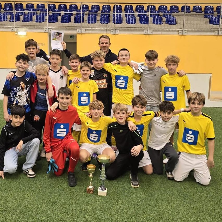 Eine Fußballmannschaft junger Jungen posiert mit einem Trainer und hält Pokale. Sie tragen gelbe und weiße Trikots mit Logo. Sie stehen auf einem grünen Feld mit leeren blauen Tribünen im Hintergrund.