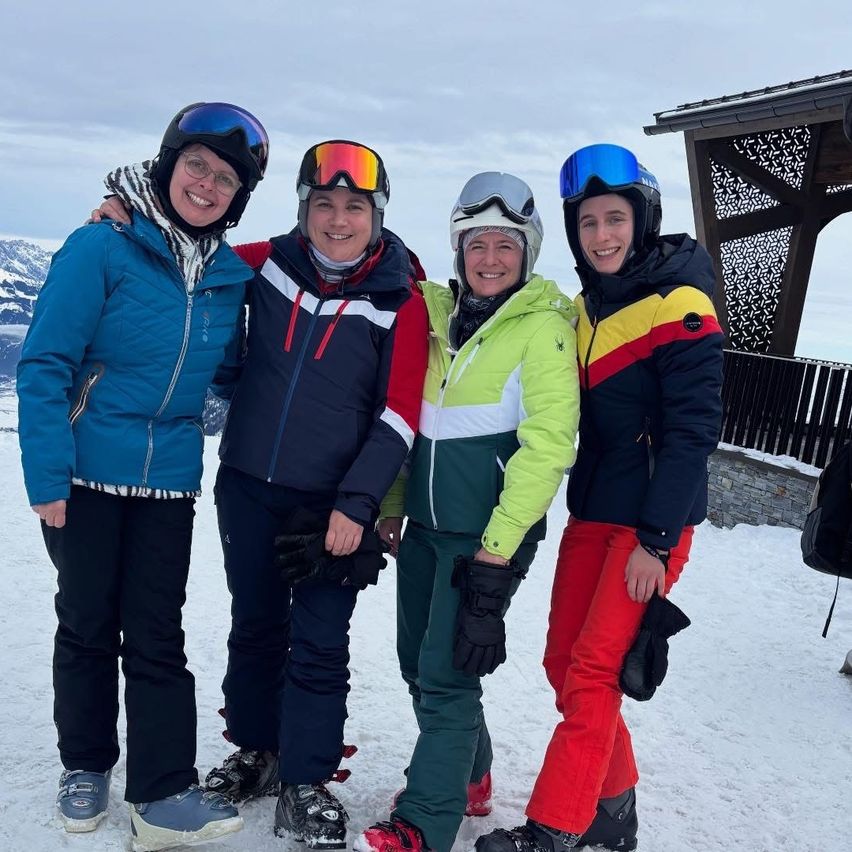 Vier Personen in Winterkleidung stehen auf einem verschneiten Berg. Sie alle lächeln und posieren für ein Foto. Hinter ihnen steht ein hölzerner Pavillon.
