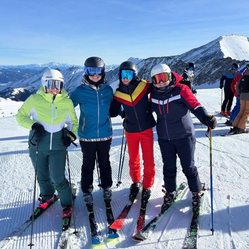 Vier Skifahrer stehen zusammen auf einem verschneiten Berg, tragen Skiausrüstung und halten Skistöcke. Sie lächeln für ein Foto mit einem verschneiten Berg im Hintergrund.