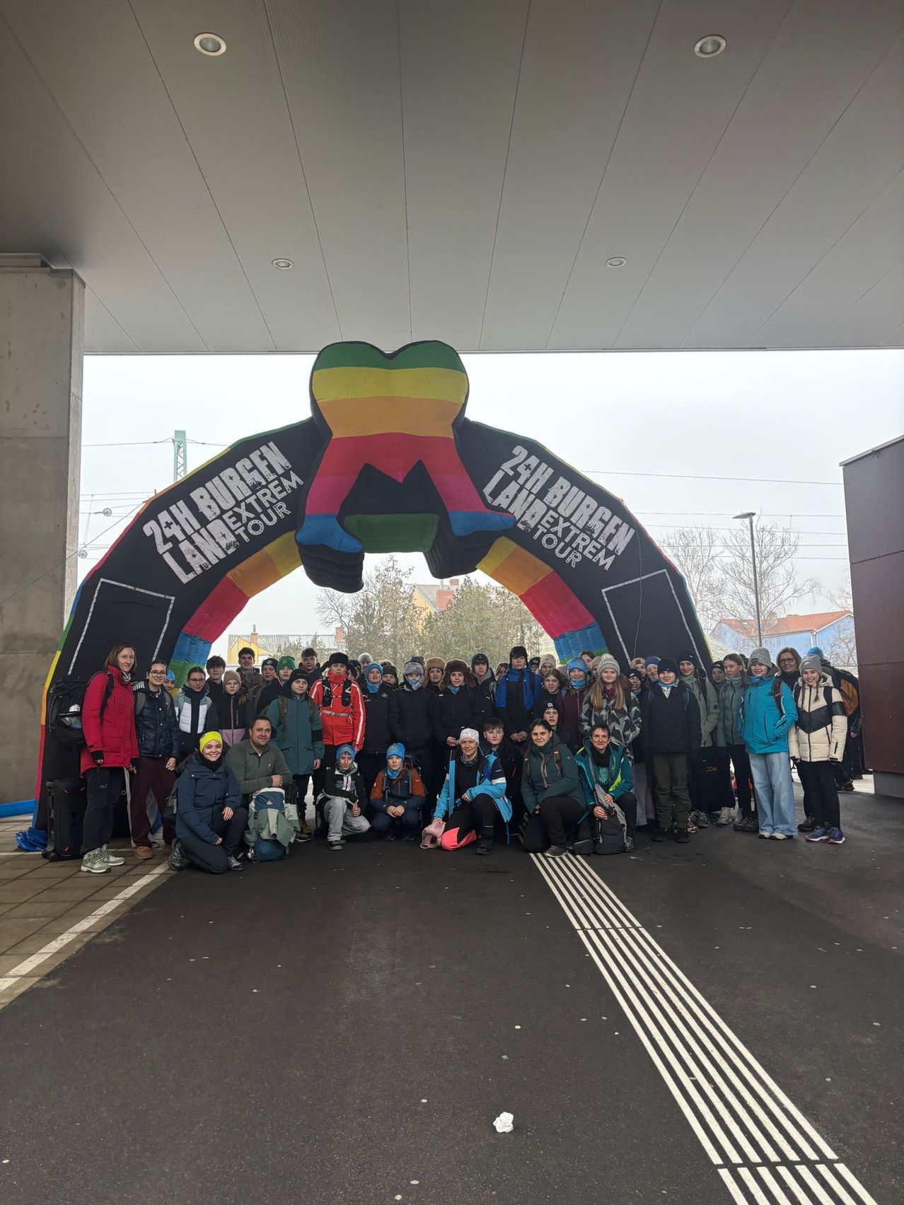 Eine Gruppe von Menschen in Winterkleidung posiert unter einem bunten Bogen mit der Aufschrift '24h Burgen Land Extrem Tour'.