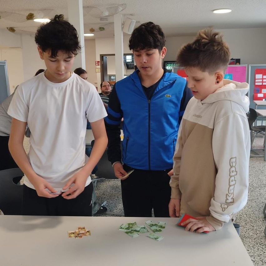 Drei Jungen sind mit Karten auf einem Tisch beschäftigt, einer hält eine rote Karte und ein anderer sieht sie an. Hinter ihnen beobachtet ein Mann in einer blauen Jacke sie.