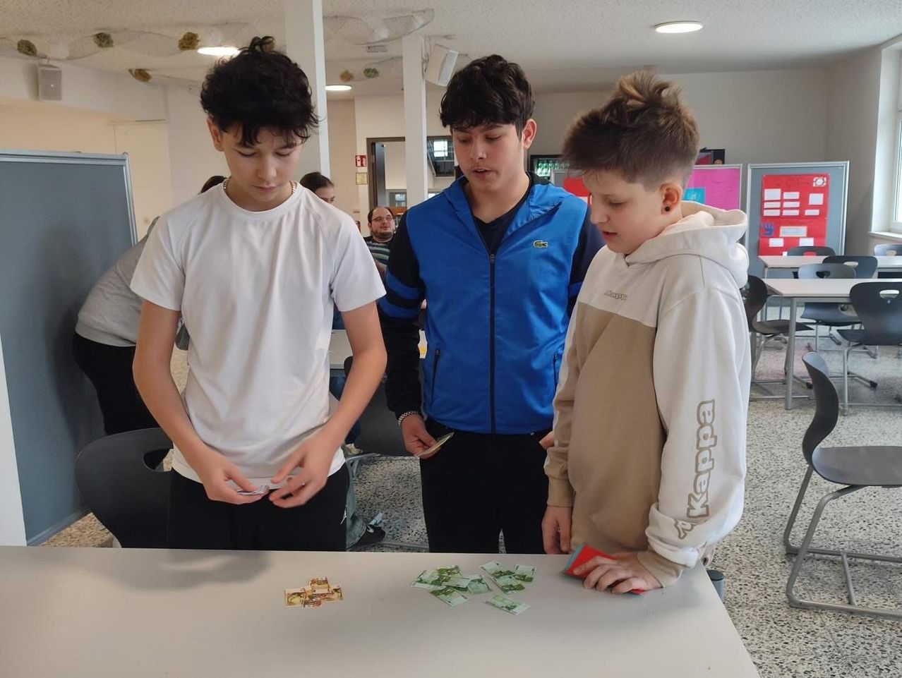 Drei Jungen sind mit Karten auf einem Tisch beschäftigt, einer hält eine rote Karte und ein anderer sieht sie an. Hinter ihnen beobachtet ein Mann in einer blauen Jacke sie.