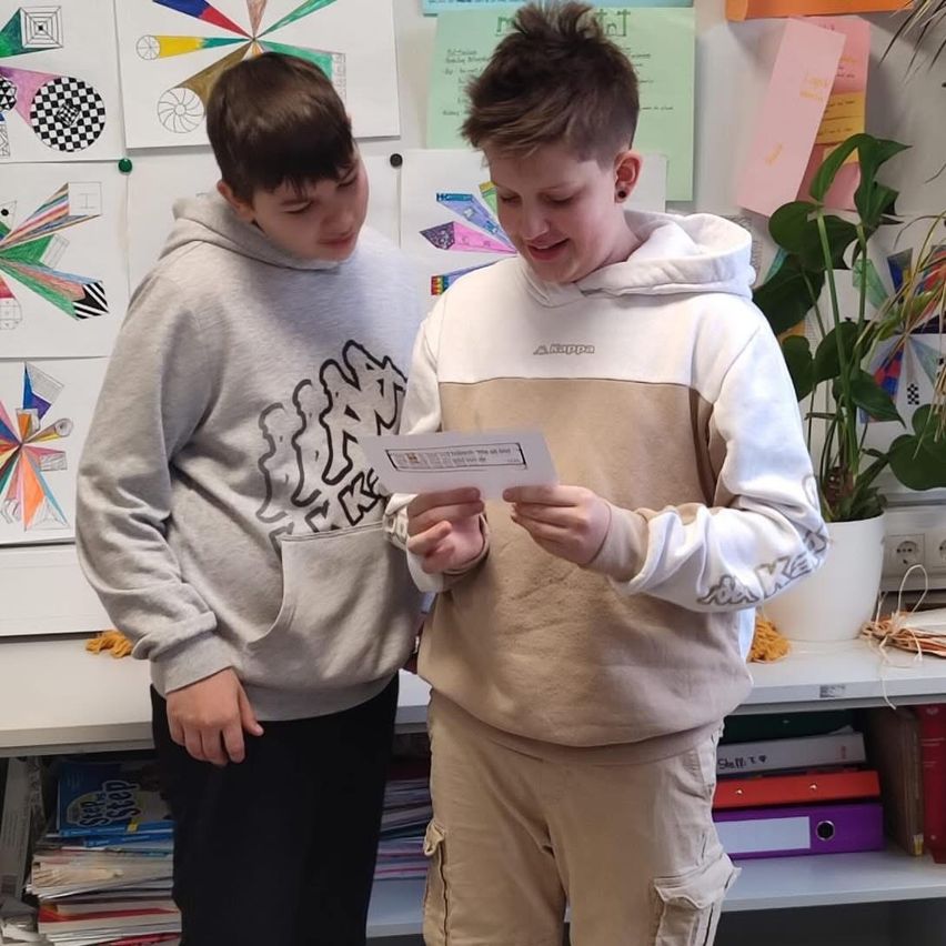 Zwei Jungen in einem Klassenzimmer, einer hält ein Papier und liest es, während der andere zusieht, mit farbenfrohen Postern an der Wand und einer Pflanze in einem weißen Topf.