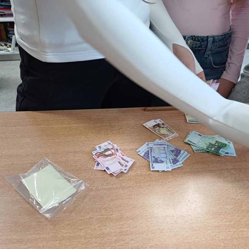 Zwei Personen stehen an einem Tisch mit Geld und einem Haftnotiz. Die Person in Weiß hält Geld, und die andere trägt Rosa und Jeans.