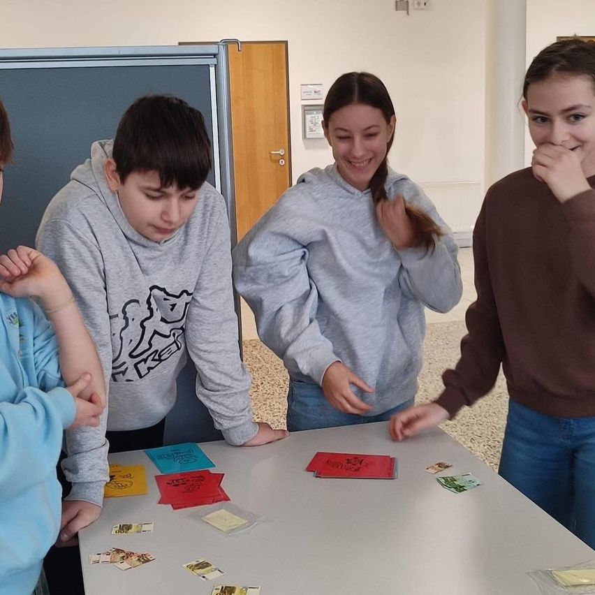 Vier Kinder sind um einen Tisch versammelt, möglicherweise beschäftigen sie sich mit einem Spiel. Auf dem Tisch befinden sich verschiedene Karten und Spielsteine. Die Kinder tragen Freizeitkleidung.
