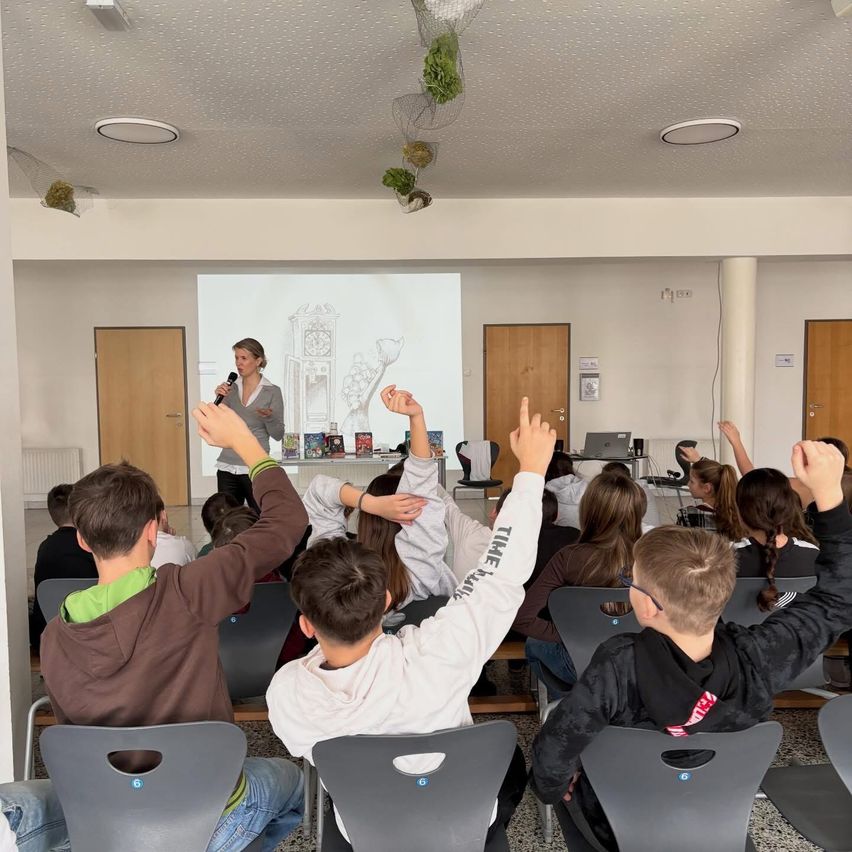 Eine Frau hält eine Präsentation für eine Klasse von Schülern ab, die ihre Hände heben und aufmerksam zuhören.