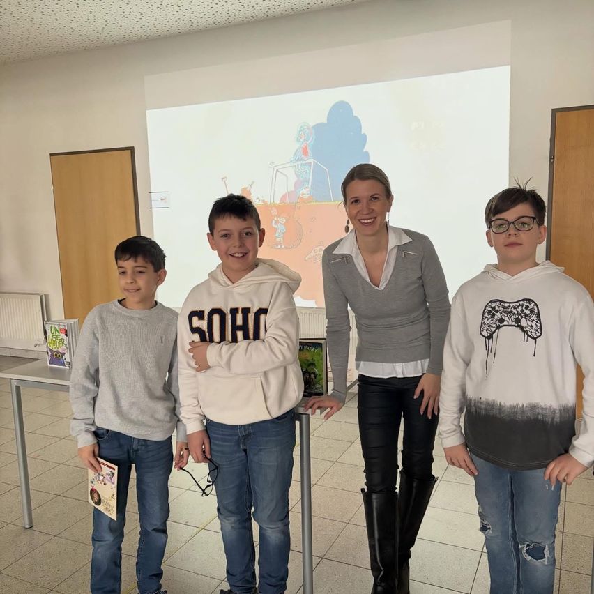 Drei Jungen und eine Frau stehen in einem Raum mit einer Projektionsleinwand. Die Jungen tragen Kapuzenpullover. Die Frau trägt ein graues Oberteil und schwarze Hose. Die Jungen lächeln.