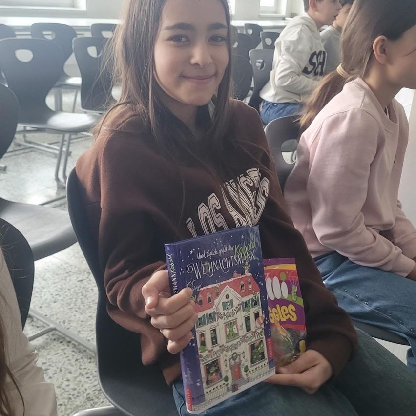 Ein Mädchen hält ein Buch mit dem Titel 'Und täglich grüßt das Karussell' und sitzt in einem Klassenzimmer mit anderen Schülern. Das Mädchen lächelt, und zwei weitere Schüler sitzen neben ihr.