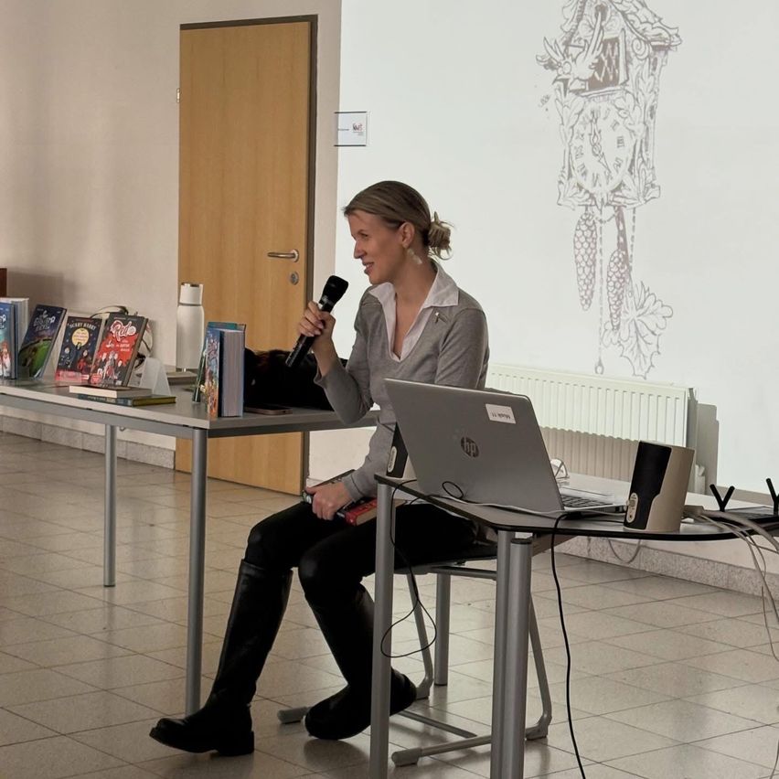 Eine Frau sitzt auf einem Stuhl und spricht in ein Mikrofon, während sie lächelt. Sie hält ein Buch auf dem Schoß. Ein Tisch hält Bücher, einen Laptop und einen Lautsprecher. Eine weiße Wand mit einem Kuckucksuhr-Zeichnung ist hinter ihr.