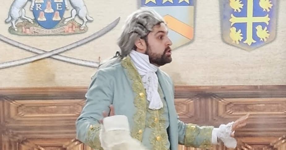 Ein Mann in einem Kostüm mit einer weißen Perücke steht und spricht, während er ein weißes Tuch hält. Hinter ihm befinden sich zwei Schwerter und Wappen an der Wand.