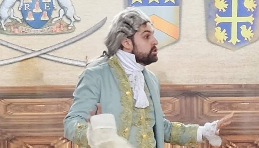Ein Mann in einem Kostüm mit einer weißen Perücke steht und spricht, während er ein weißes Tuch hält. Hinter ihm befinden sich zwei Schwerter und Wappen an der Wand.