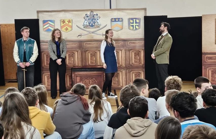 Fünf Personen, drei Frauen und zwei Männer, stehen auf der Bühne und führen vor einem großen Publikum in einem Saal auf.