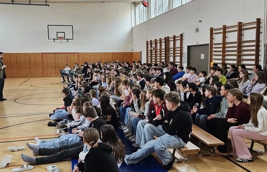 Eine große Gruppe von Schülern sitzt auf Bänken in einer Turnhalle und beobachtet etwas. Einige Schüler sind auf dem Boden, während andere sitzen. Die Turnhalle hat Holzwände und einen Basketballkorb.