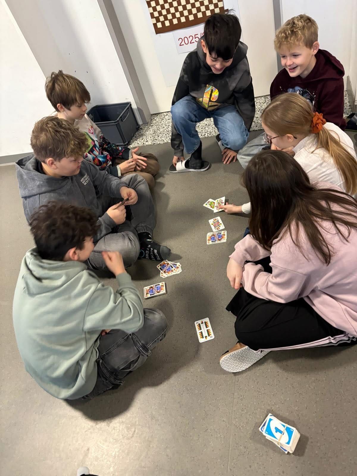 Sechs Kinder sitzen im Kreis auf einem gefliesten Boden und spielen ein Kartenspiel. Einige Karten liegen auf dem Boden. Ein Kind schaut sich die Karten an, während ein anderes lächelt.