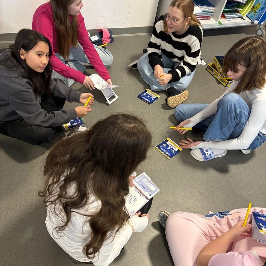 Sechs Mädchen sitzen auf dem Boden und sind in eine Aktivität vertieft. Jedes hält einen Stift und Papier. Sie scheinen ein Rätsel oder eine Aktivität zu machen.