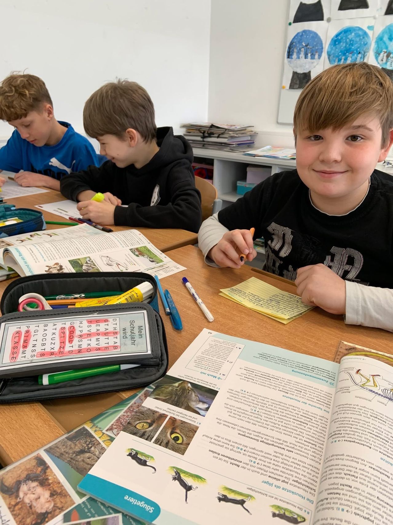 Drei Jungen im Klassenzimmer, einer lächelt in die Kamera, die anderen beiden schauen auf ihre Papiere. Schulmaterial liegt auf dem Schreibtisch.