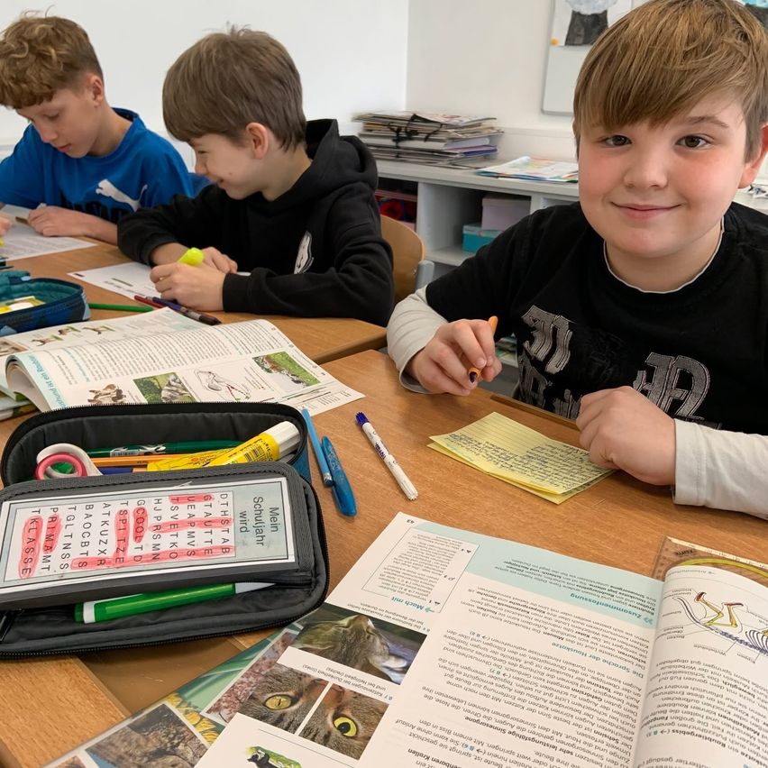 Drei Jungen im Klassenzimmer, einer lächelt in die Kamera, die anderen beiden schauen auf ihre Papiere. Schulmaterial liegt auf dem Schreibtisch.
