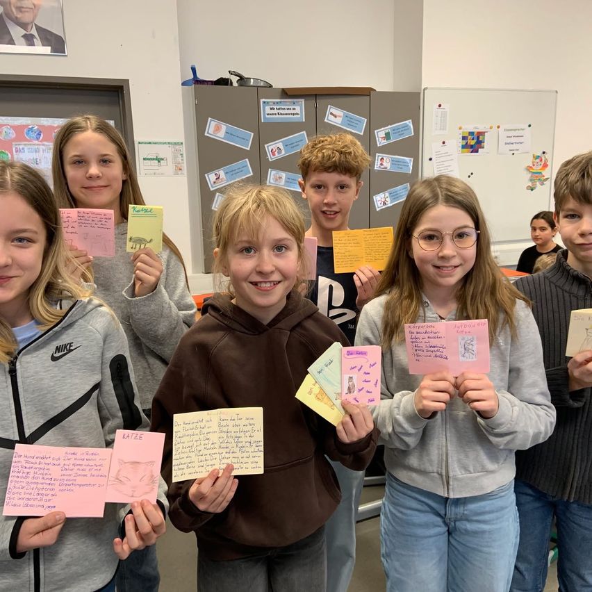 Gruppe lächelnder Kinder in einem Klassenzimmer, die bunte Karten mit Zeichnungen und Text hochhalten, mit einer Tafel und einem Bulletin Board im Hintergrund.