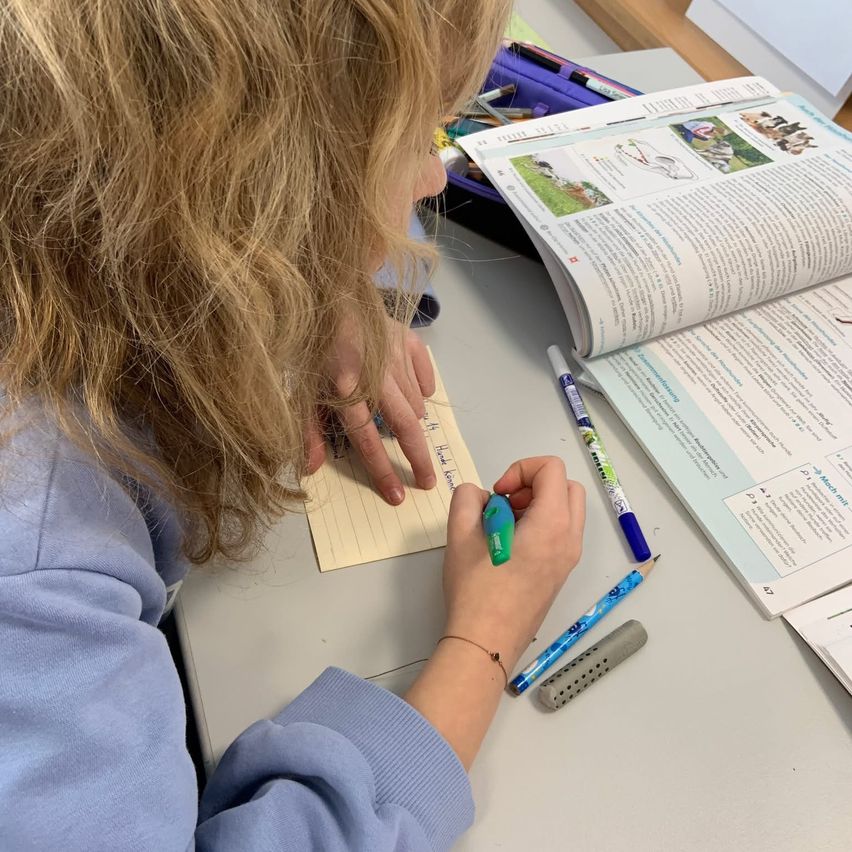 Eine blonde Schülerin schreibt mit einem Marker auf einem Zettel, während sie am Schreibtisch sitzt. Auf dem Tisch liegen Bücher, ein Bleistift und ein Spitzer.
