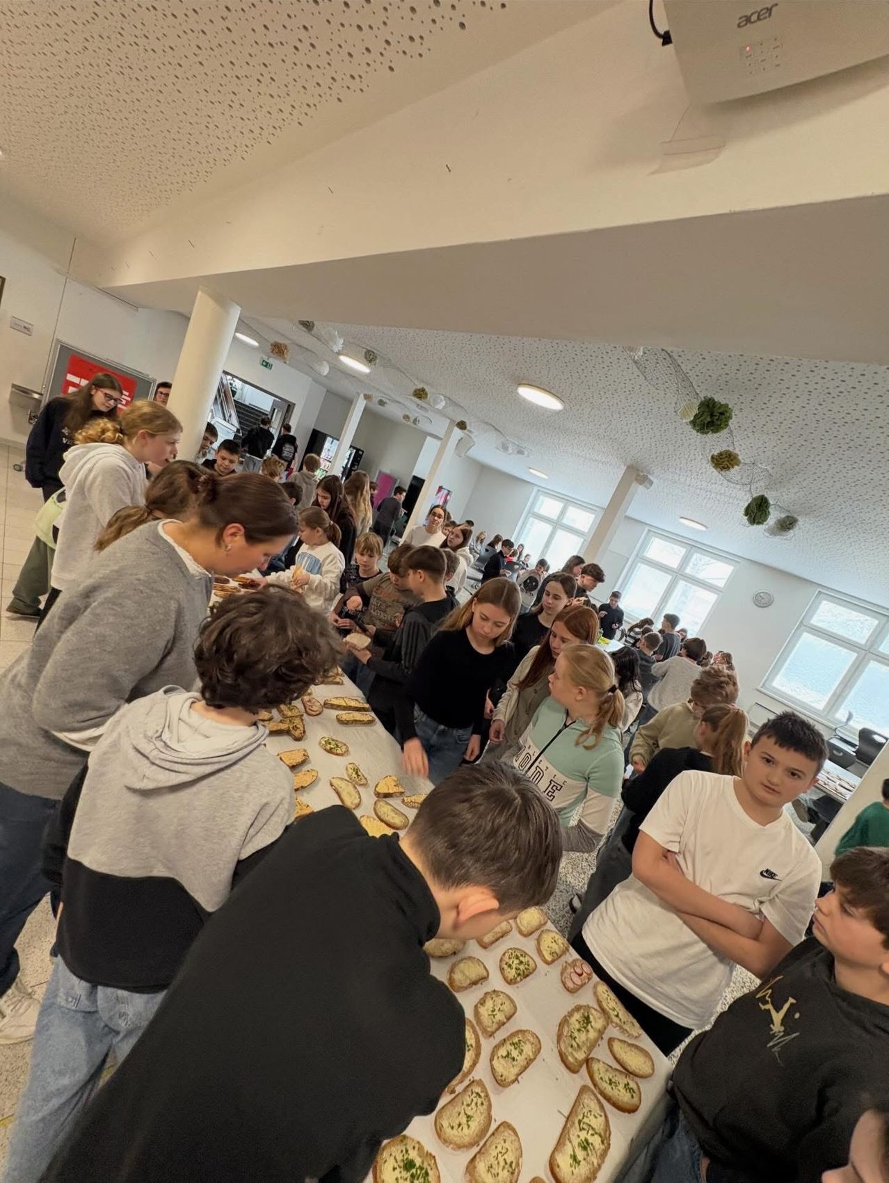 Viele Kinder sind um einen Tisch in einem Raum versammelt und essen Snacks. Der Tisch ist mit verschiedenen Speisen gefüllt. Einige Kinder schauen sich das Essen an. Der Raum hat Fenster und weiße Wände.