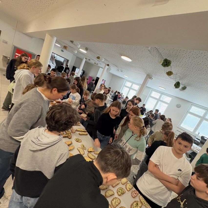 Viele Kinder sind um einen Tisch in einem Raum versammelt und essen Snacks. Der Tisch ist mit verschiedenen Speisen gefüllt. Einige Kinder schauen sich das Essen an. Der Raum hat Fenster und weiße Wände.