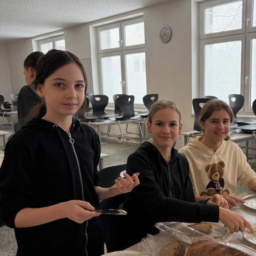 Drei junge Mädchen lächeln in einem Klassenzimmer, zwei halten ein Stück Brot. Hinter ihnen sind Stühle in Reihen aufgestellt, und eine Uhr ist an der Wand montiert. Sonnenlicht filtert durch die Fenster.