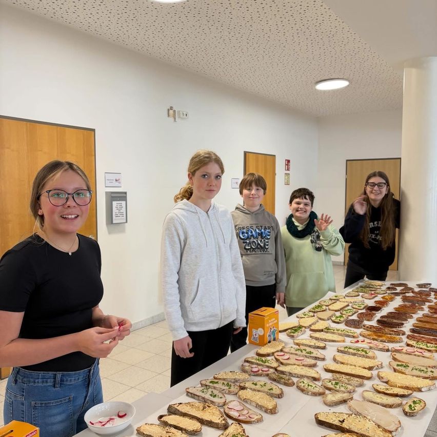 Fünf junge Leute stehen hinter einem Tisch mit Sandwiches. Der Tisch ist voll mit verschiedenen Arten von Sandwiches. Der Raum hat weiße Wände und eine Decke mit Lichtern.