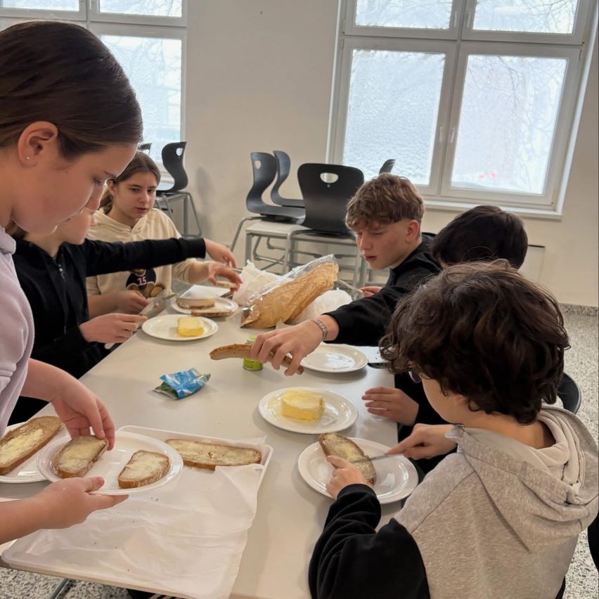 Eine Gruppe junger Schüler sitzt um einen Tisch herum und isst Sandwiches. Der Tisch ist mit Tellern mit Brot, Butter und Sandwiches bedeckt. Eine Frau steht links, möglicherweise beim Servieren. Der Raum hat große Fenster.