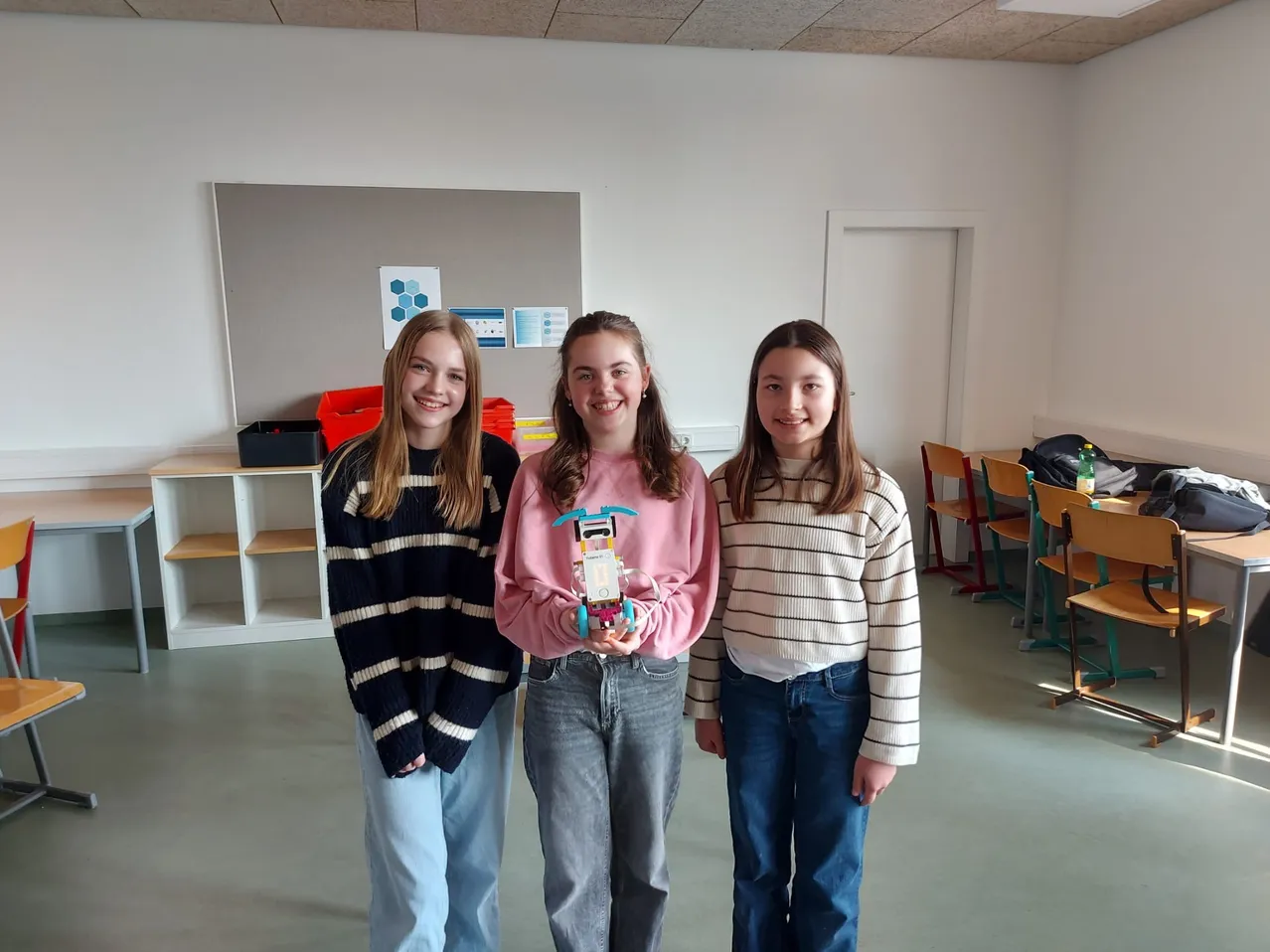 Drei junge Mädchen stehen in einem Raum mit einer grauen Wand und einem weißen Regal. Das Mädchen in der Mitte hält ein Spielzeug. Sie lächeln und posieren für ein Foto.